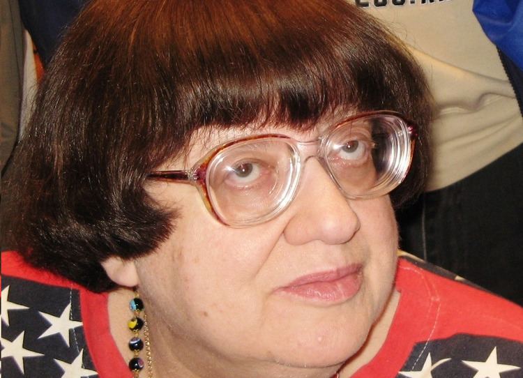 Valeriya Novodvorskaya seriously looking from above with a short black hair with bangs, wearing an eyeglass, a bead necklace, and a black and orange blouse with star prints