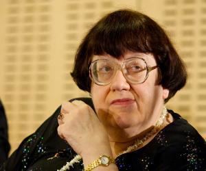 Valeriya Novodvorskaya seriously looking at someone with a short black hair with bangs, wearing an eyeglass, a gold watch on her left wrist, a white bead necklace, and a black top