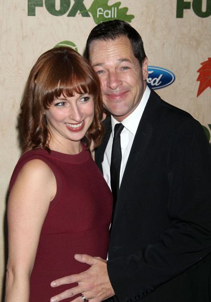 Vanessa Claire Stewart smiling while French Stewart with a tight-lipped smile while holding his wife's tummy. Vanessa with blonde hair and wearing a maroon sleeveless dress while French wearing a black coat over white long sleeves, and a black necktie.