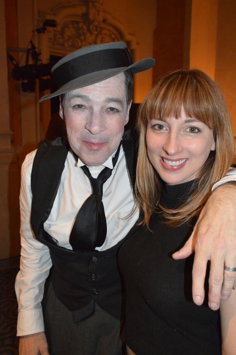Vanessa Claire Stewart smiling while French Stewart with a tight-lipped smile while his arm on the shoulder of his wife. Vanessa with blonde hair and wearing a black knitted crop top while French wearing a black and gray cap, black vest, white long sleeves, black tie, and gray pants.
