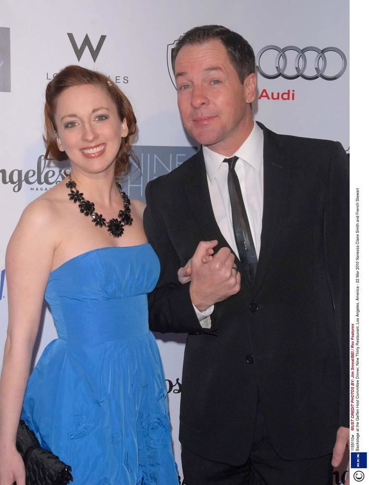 Vanessa Claire and French Stewart smiling while holding hands. Vanessa with blonde hair, wearing a necklace and blue tube dress while French wearing a black coat over white long sleeves, black pants, and a black necktie.