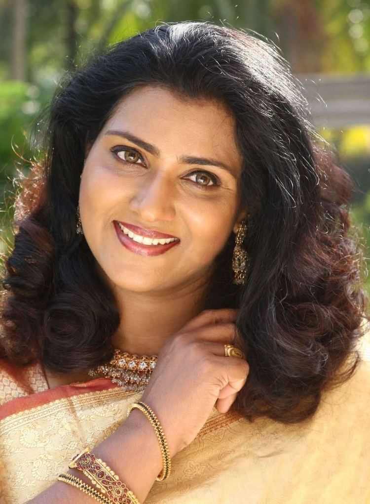 Vani Viswanath smiling, with wavy hair, wearing earrings, a necklace, bracelets, and traditional Indian clothing for women.