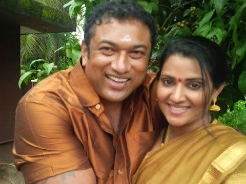 Vani Viswanath smiling and wearing a brown dress together with his husband Baburaj wearing a brown polo shirt with a smiling face.