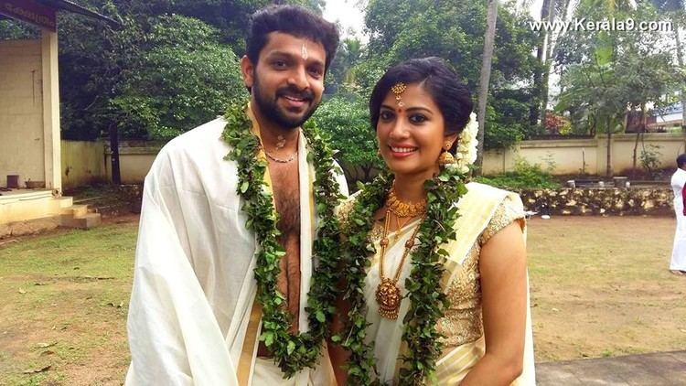 Murali Krishna smiling with Shivada Nair at their wedding reception while wearing a white and gold dress, garlands, and some jewelry