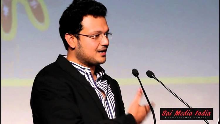 Varun Manian speaking on a podium and wearing a blue and white striped shirt under a black coat and eyeglasses
