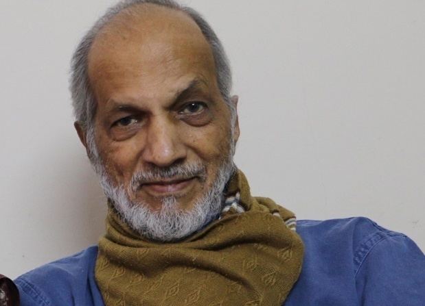 Vasant Gowarikar with a tight-lipped smile, mustache, and beard while wearing a brown scarf and blue polo