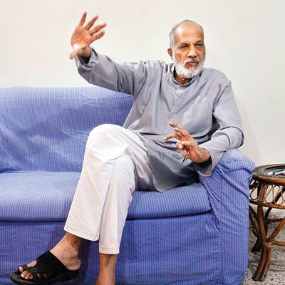 Vasant Gowarikar talking to someone with hand gestures while sitting on a blue couch and wearing gray long sleeves and white pants