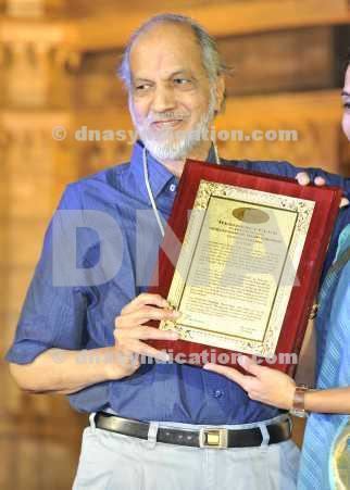 Vasant Gowarikar smiling while holding a certificate while wearing a blue polo, black belt, and white pants