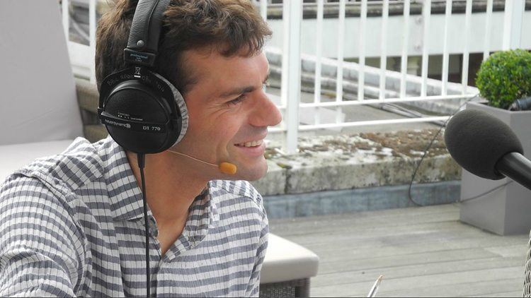 Vassos Alexander smiling while wearing a headphone and white and grey long sleeves