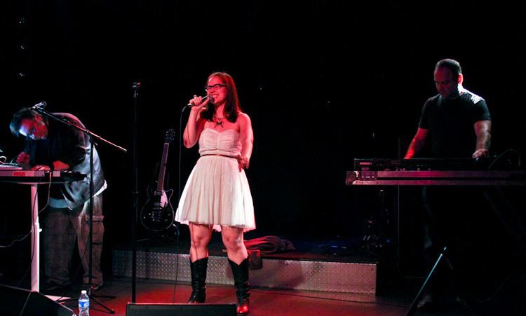 The Venus Hum performing on stage, an electronic pop music group with the members: Kip Kubin, Annette Strean, and Tony Miracle (from left to right). Kip and Tony are wearing black shirts while Annette is wearing eyeglasses, a necklace, a white tube dress, and black boots.