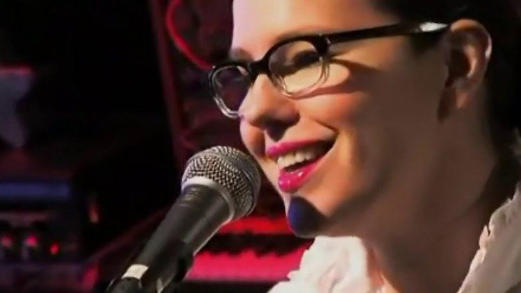 Annette Strean is smiling while singing, wearing eyeglasses and a white top.