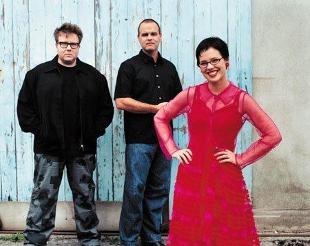 Members of the Venus Hum: Kip Kubin, Tony Miracle, and Annette Strean (from left to right). Kip is wearing eyeglasses, a black jacket, and camouflage pants while Tony is wearing a black polo shirt and blue pants and Annette is wearing eyeglasses and a red dress.