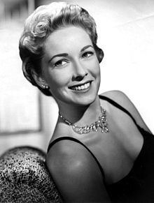 Vera Miles smiling and sitting on a couch with short curly hair while wearing a black dress and necklace