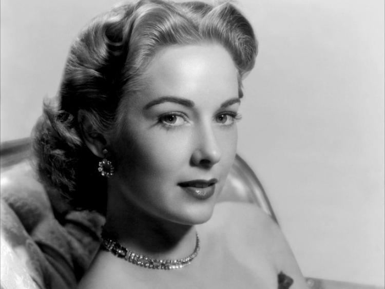 Vera Miles smiling and sitting on a couch while wearing a black tube dress with necklace and earrings