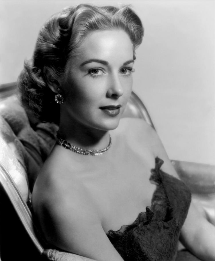 Vera Miles smiling and sitting on a couch while wearing a tube dress with a necklace and earrings
