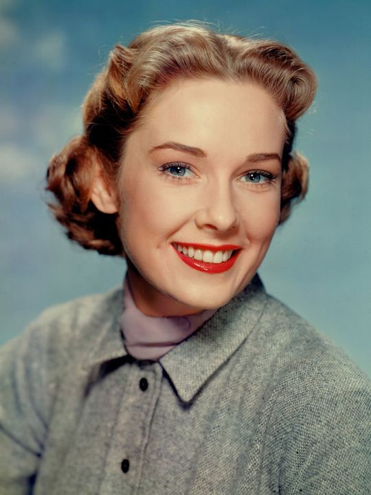 Vera Miles smiling with short curly blonde hair while wearing a gray buttoned blouse