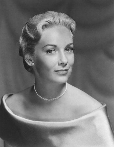 Vera Miles smiling with short curly hair while wearing an off-shoulder dress paired with pearl earrings and necklace