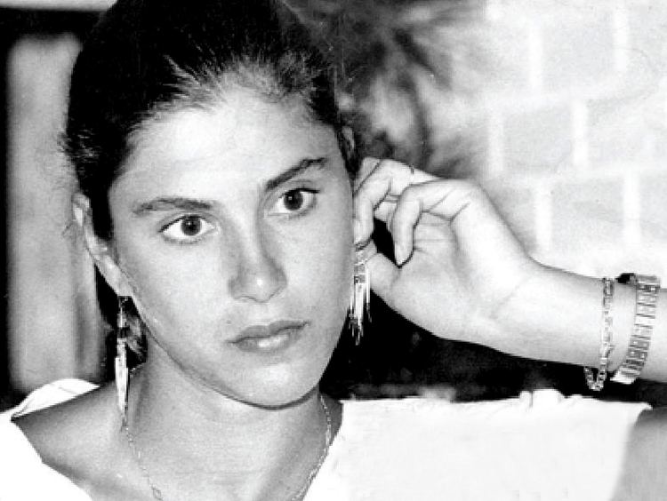 Vera Mossa holding her earrings while looking at something and wearing a t-shirt, necklace, bracelet, and wristwatch
