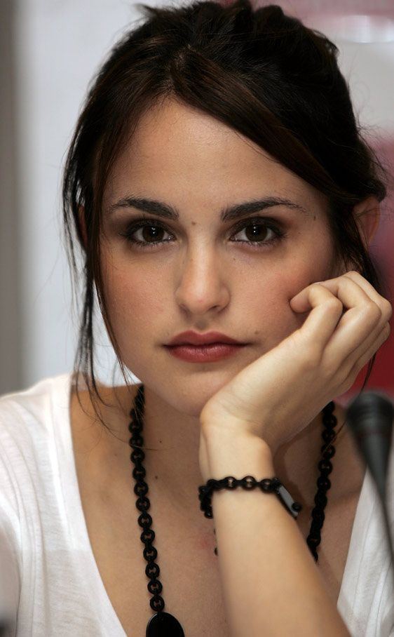 Verónica Echegui with a neutral face and hands holding her face in front of a mic, is a Spanish actress. She has dark brown hair in a ponytail with side bangs, a black beaded bracelet on her left wrist, a lengthy black beaded necklace, and wearing a white V-neck shirt.