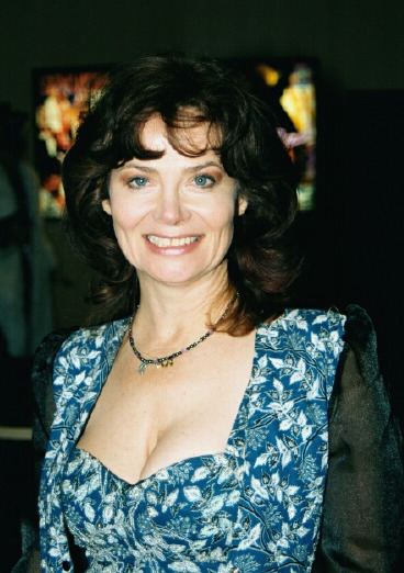 Veronica Hart smiling with black curly hair and blue eyes while wearing a necklace and a white and blue blouse with black long sleeves and a sweetheart neckline that exposes her cleavage