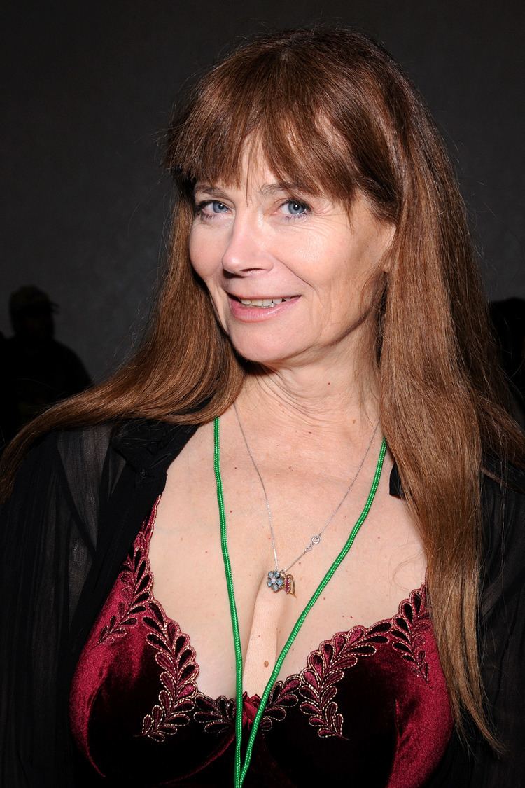 Veronica Hart smiling with brown hair and blue eyes while wearing a necklace, green lanyard, black blazer, and a maroon blouse with a low-cut neckline that exposes her cleavage