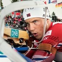 Vetle Sjåstad Christiansen looking serious holding a biathlon rifle with DNB print on it, wearing a white head cap with a small Norwegian flag, and a printed red snowsuit.