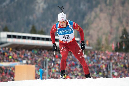 Vetle Sjåstad Christiansen looking down while on his ski, holding his ski poles with a crowd in the background. He is wearing a white head cap and an action camera, ski goggles, black gloves, with his athlete number 42 on his red snowsuit.