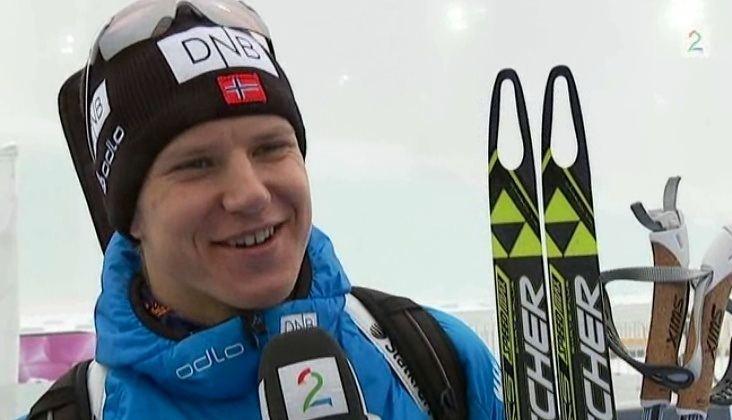 Vetle Sjåstad Christiansen smiling while having an interview, holding his skis and ski poles, is a Norwegian biathlete, wearing a ski goggles on his black ski bonnet with a small Norwegian flag, and a blue insulated jacket with prints on.