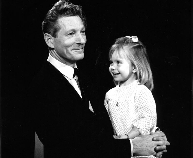 Young Victoria Paige Meyerink (right) smiling and sitting on Danny Kaye’s (left) lap in an old photograph. Victoria has blonde hair with a ribbon clip, wearing a necklace, bracelet, and a white long sleeve dress, while Danny has black hair, wearing a white polo with a black necktie and a black coat