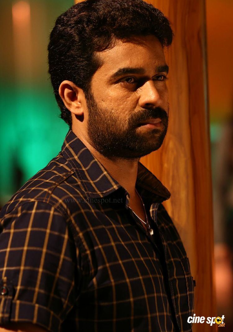 Vijay Babu looking afar with mustache and beard while wearing a brown and cream checkered polo