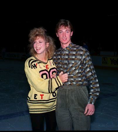 Vikki Moss (left) smiling and grabbing Wayne Gretzky (right) long sleeve.  Vikki has blonde kinky hair, wearing an earring, yellow knitted long sleeve and black pants while Wayne smiling while his right arm around Vikki’s shoulders, he has black hair, wearing a tuck in stripes long sleeve polo and gray pants
