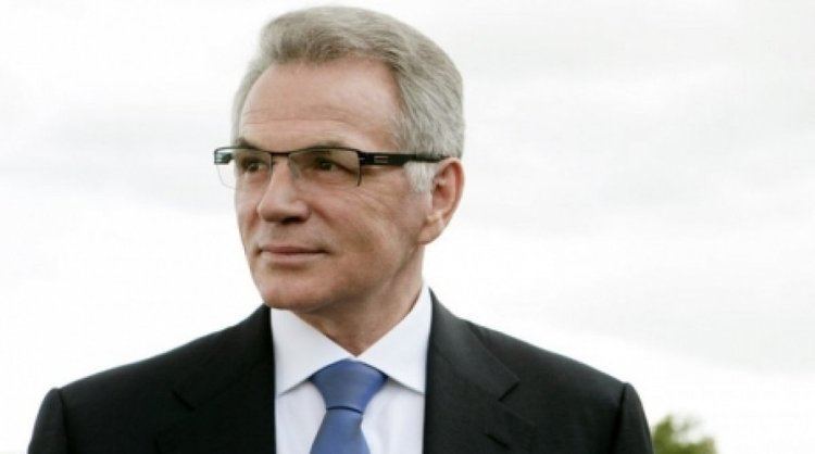 Viktor Khrapunov with a tight-lipped smile while wearing eyeglasses, a black coat, white long sleeves, and blue necktie