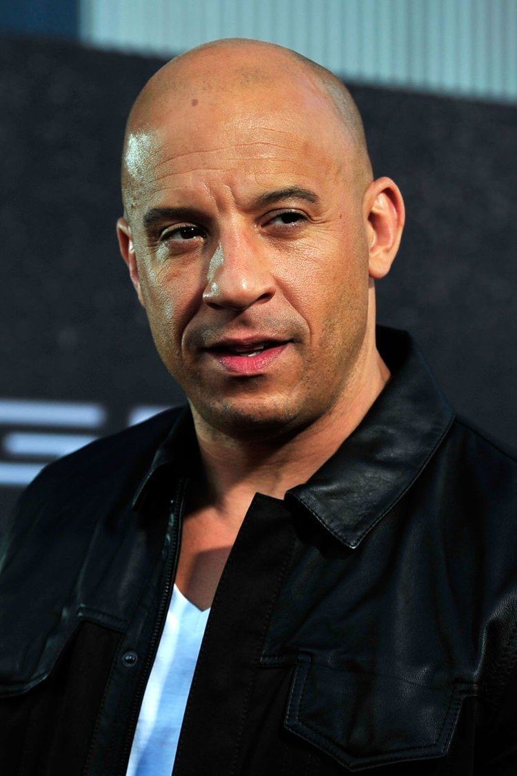 Vin Diesel smiling with a bit of facial hair and wearing a white shirt underneath a black leather jacket.