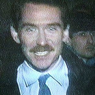Vincent Hanley smiling having a mustache and a person behind him while wearing a white shirt under a black coat and a blue necktie
