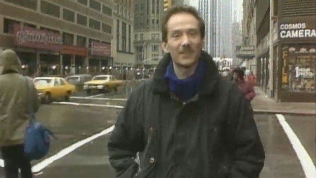 Vincent Hanley reporting on the side of the street with yellow cabs, people and buildings in the background and wearing a black coat and a blue scarf around his neck