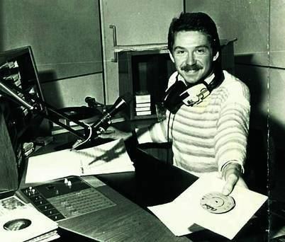 Vincent Hanley smiling with a mustache and handing a CD inside the radio station with a headset around his neck and wearing a striped shirt