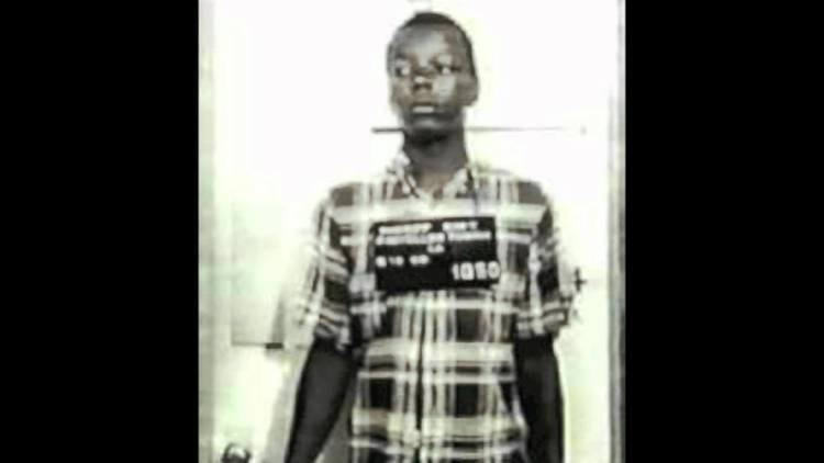 Vincent Simmons standing while looking sideways was a life prisoner at Angola State Prison in Louisiana, where he was sentenced to 100 years in July 1977. He is wearing a tag over his neck with writings on it, and a collared checkered 3/4 sleeve shirt.