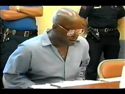 Vincent Simmons sitting and talking while looking down, was a life prisoner at Angola State Prison in Louisiana, during his meeting with the Sanders Twins in 1999. There are two policemen with guns in their holsters in the background and a table and papers in front. Vincent has a bald head, an airplane-pendant necklace, and sunglasses while wearing a light blue collared long sleeve.