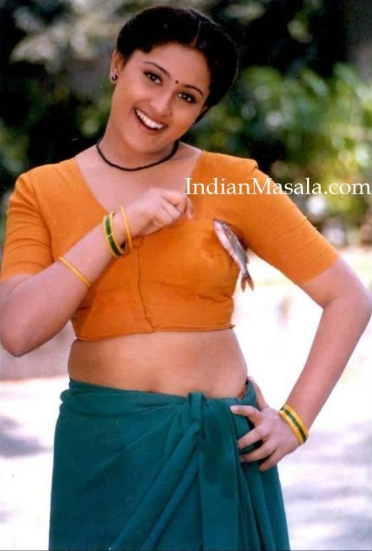 Vineetha is smiling while holding a tie of fish on her left breast, wearing earrings, a necklace, green and yellow bracelets, an orange blouse, and a green skirt.