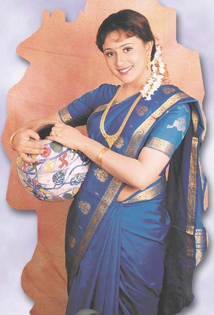 Vineetha is smiling while holding a colorful jar on her waist, wearing a floral headdress, gold earrings, gold necklace, gold bangles, gold rings, and a blue and golden saree dress.