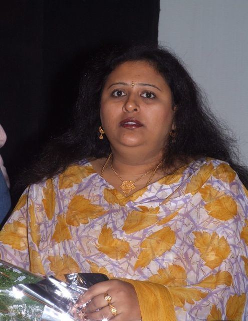 Vineetha with a serious face while holding a silver gift wrapper, with messy hair, wearing gold earrings, gold necklaces, gold rings, and a yellow and white top.