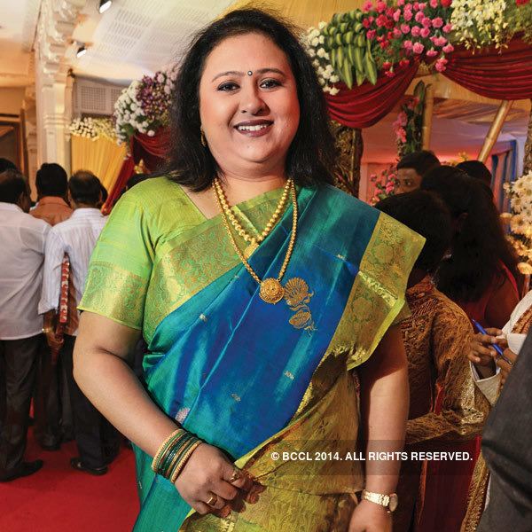 Vineetha is smiling with people at the back, with messy hair, wearing a gold necklace, bangles, rings, a watch, and a green and blue saree.