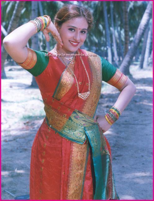 Vineetha is smiling while her hands are on her face and hip with coconut trees in the background, wearing gold necklaces, colorful bangles, and a multi-colored saree dress.