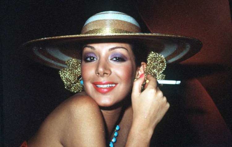 Virginia Vallejo smiling while holding a cigarette and wearing hat, gold earrings and blue necklace