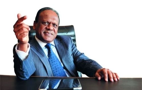 Vivian Reddy smiling while sitting on the chair and wearing a striped coat, white long sleeves, and blue necktie