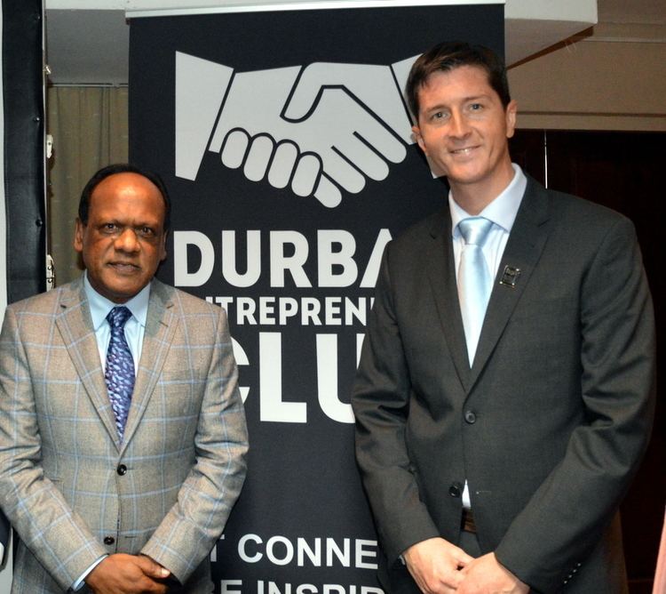 Vivian Reddy smiling and wearing a checkered coat, light blue long sleeves, and blue necktie while Grant Gavins standing next to him