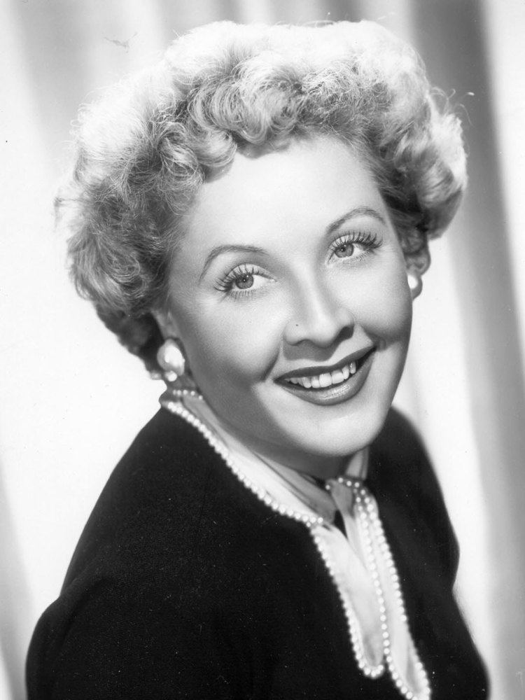 Vivian Vance is smiling, has short curly hair, wearing pearl earrings and a black and white top with a pearl strand design.