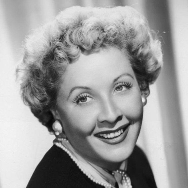 Vivian Vance is smiling, has short curly hair, wearing pearl earrings and a black and white top with a pearl strand design.