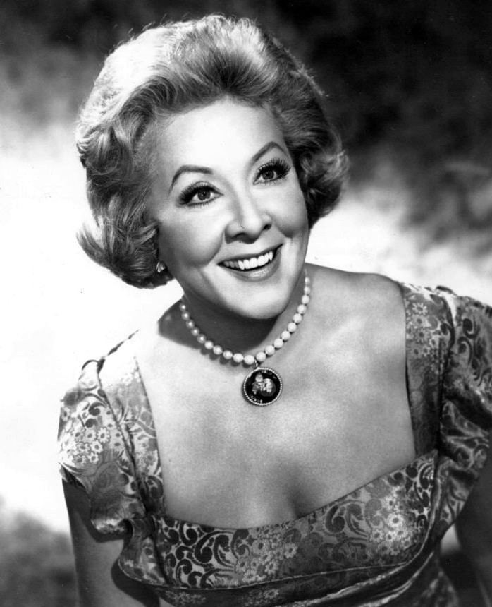 Vivian Vance is smiling, has short curly hair, wears earrings and a white pearl necklace with a black pendant, and wears a floral cleavage showing dress.