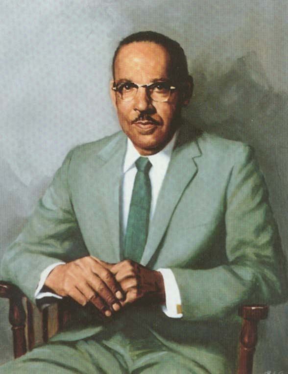 Vivien Thomas sitting on a wooden chair while holding his hands together with a mustache, wearing an eyeglass and a white long sleeve with a dark green necktie under his light green coat